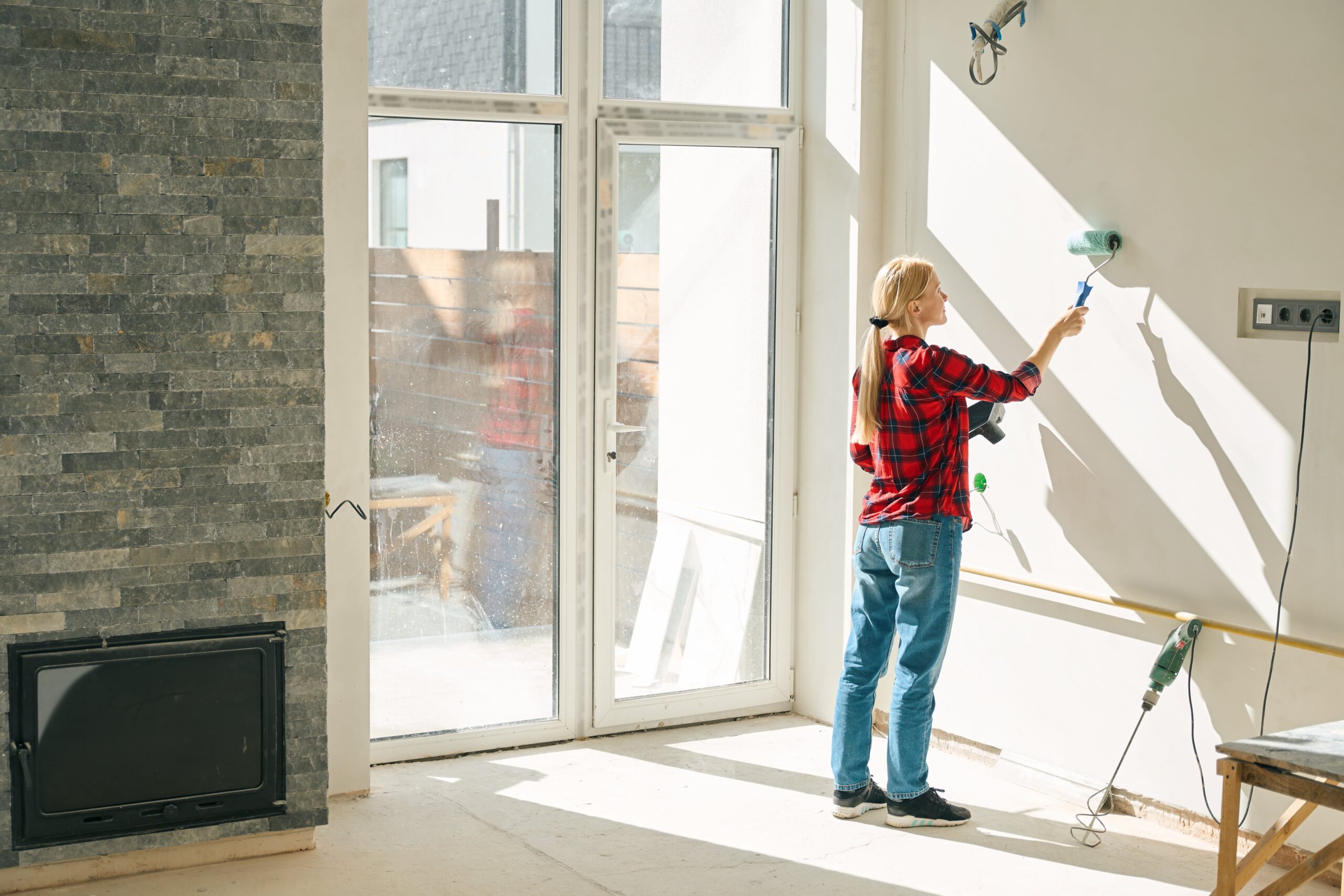 A back view of a woman applying paint to the walls of a room in a house with a roller brush.