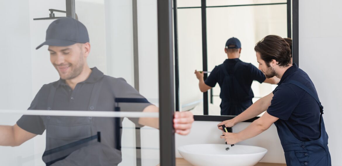 Two professional handymen installing a modern glass shower cabin and a white ceramic sink in a bathroom. One handyman, smiling and wearing a cap, is measuring the shower door, while the other is adjusting the faucet on the sink.