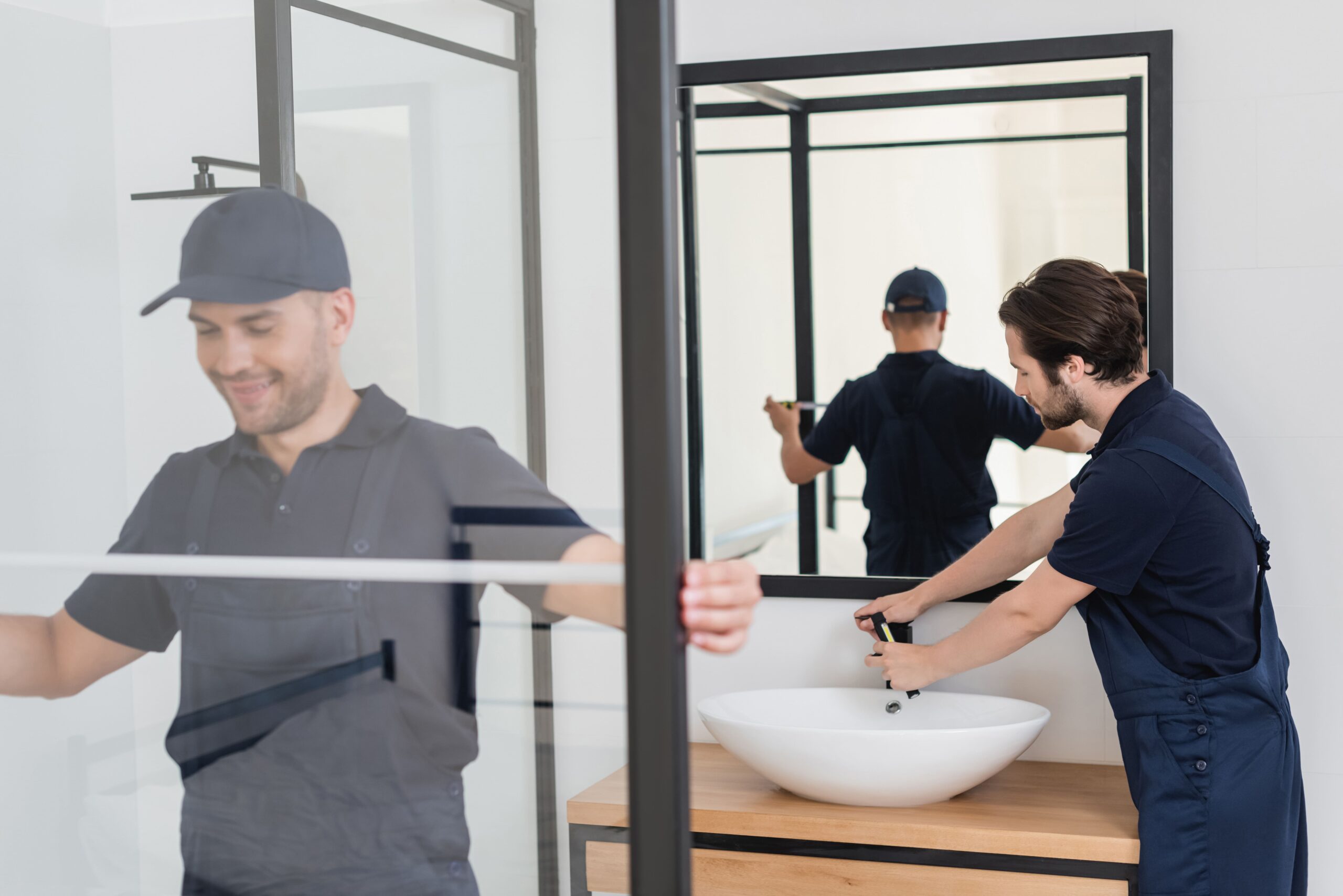 Two professional handymen installing a modern glass shower cabin and a white ceramic sink in a bathroom. One handyman, smiling and wearing a cap, is measuring the shower door, while the other is adjusting the faucet on the sink.