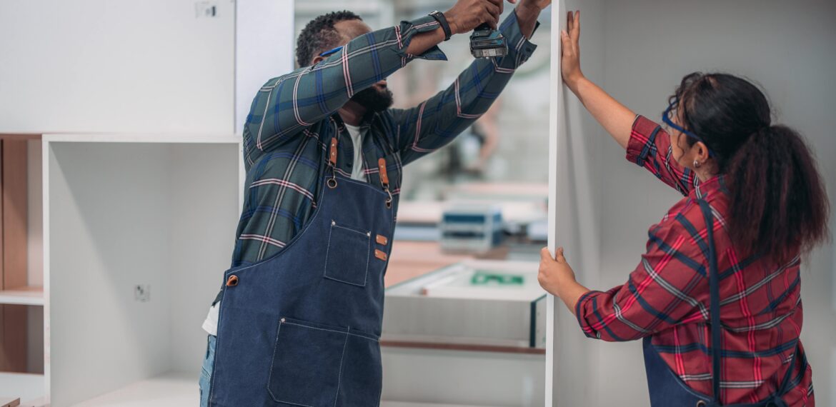 A carpenter wearing a working apron uses a drilling machine to fix a cabinet while a woman carpenter is assisting him.