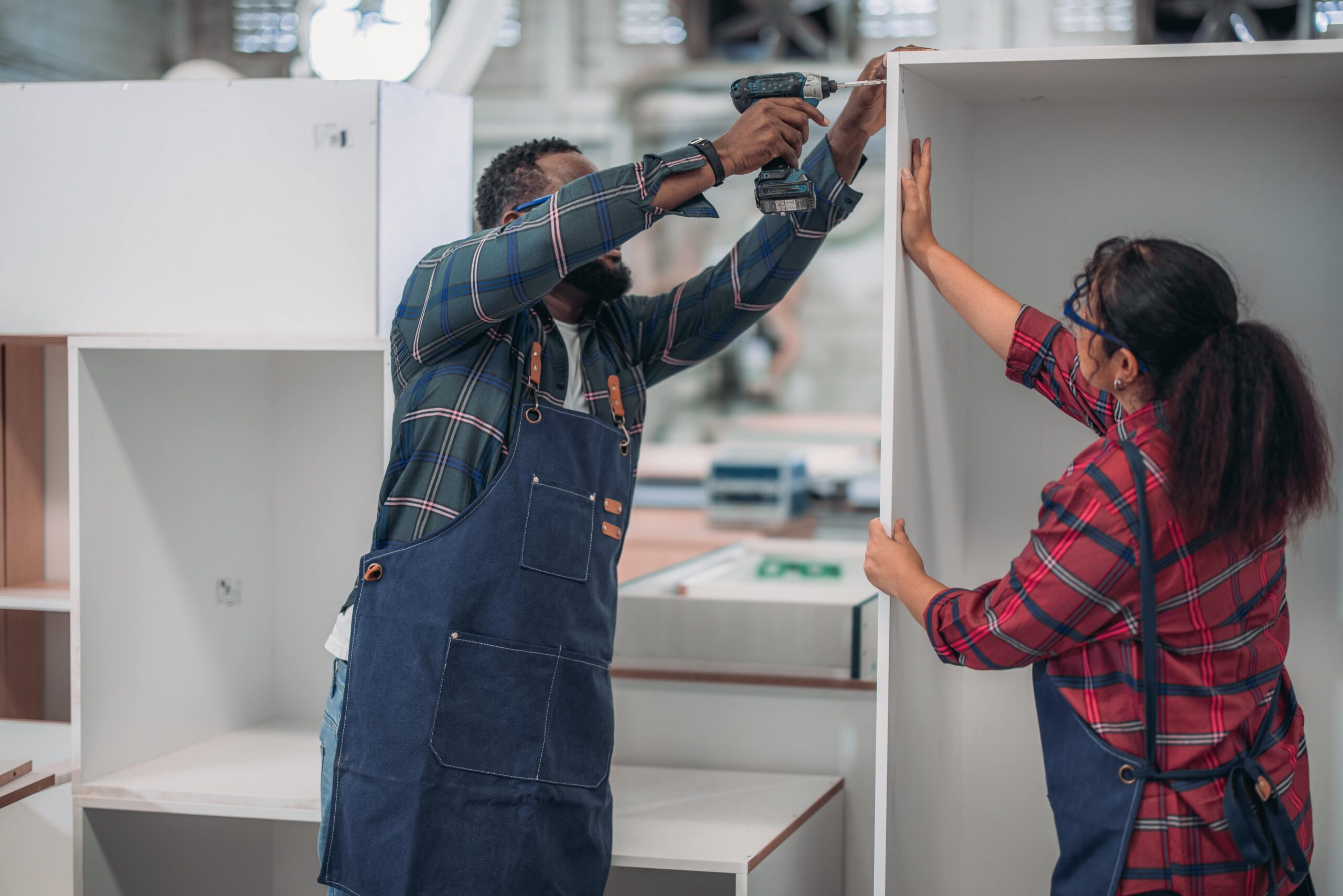 A carpenter wearing a working apron uses a drilling machine to fix a cabinet while a woman carpenter is assisting him.