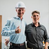 A home remodeler wearing a hard hat smiles at the camera along with a male client.